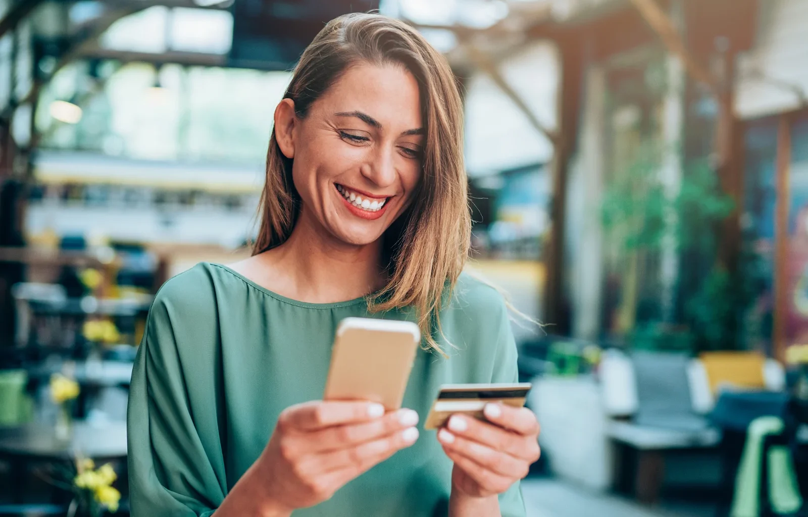 lächelnde Frau hält eine Kreditkarte in der einen und ihr iPhone in der anderen Hand