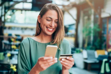 lächelnde Frau hält eine Kreditkarte in der einen und ihr iPhone in der anderen Hand