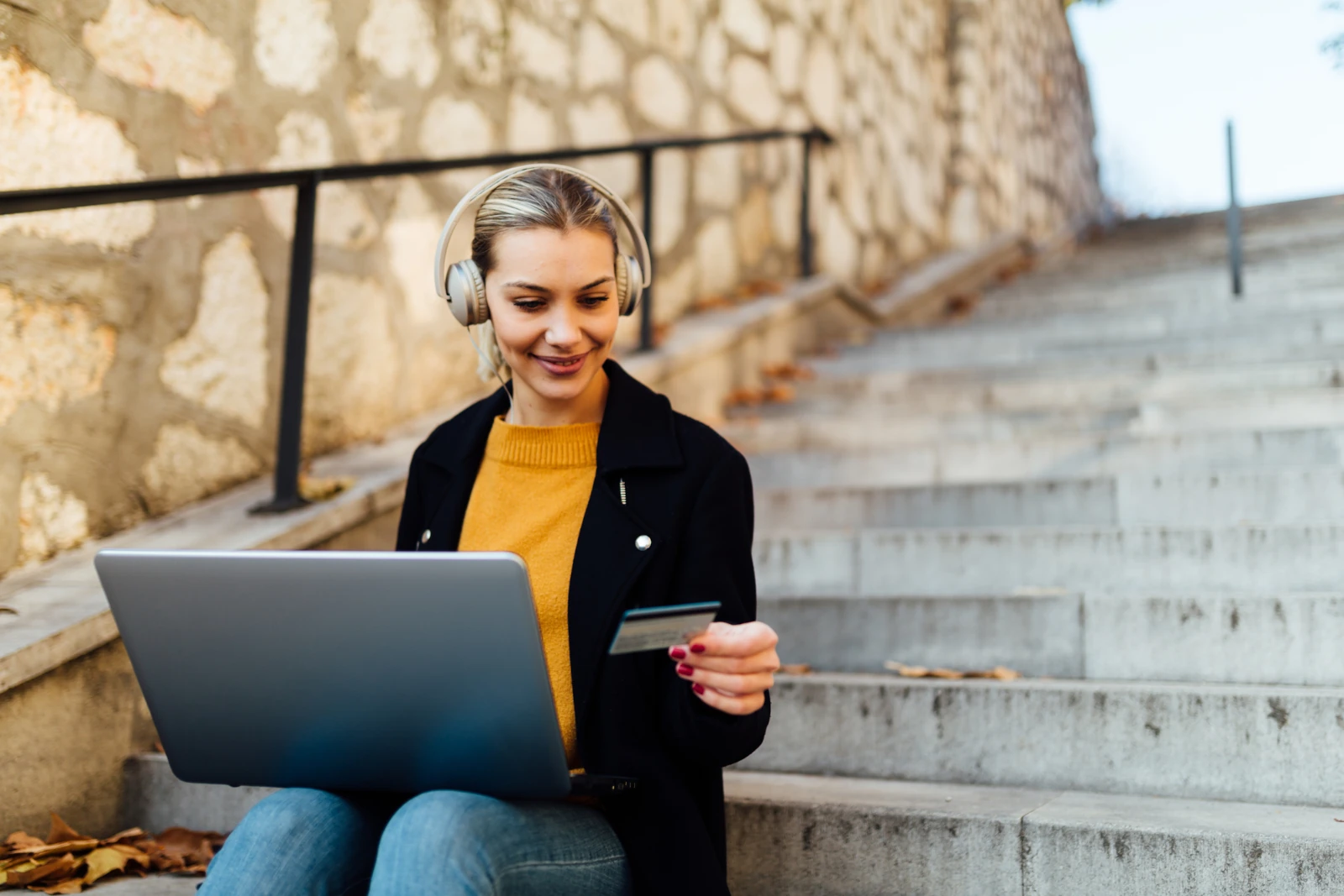Junge Frau beim Online-Shopping mit Kreditkarte und Laptop sitzt auf Stufen im Freien in der Stadt