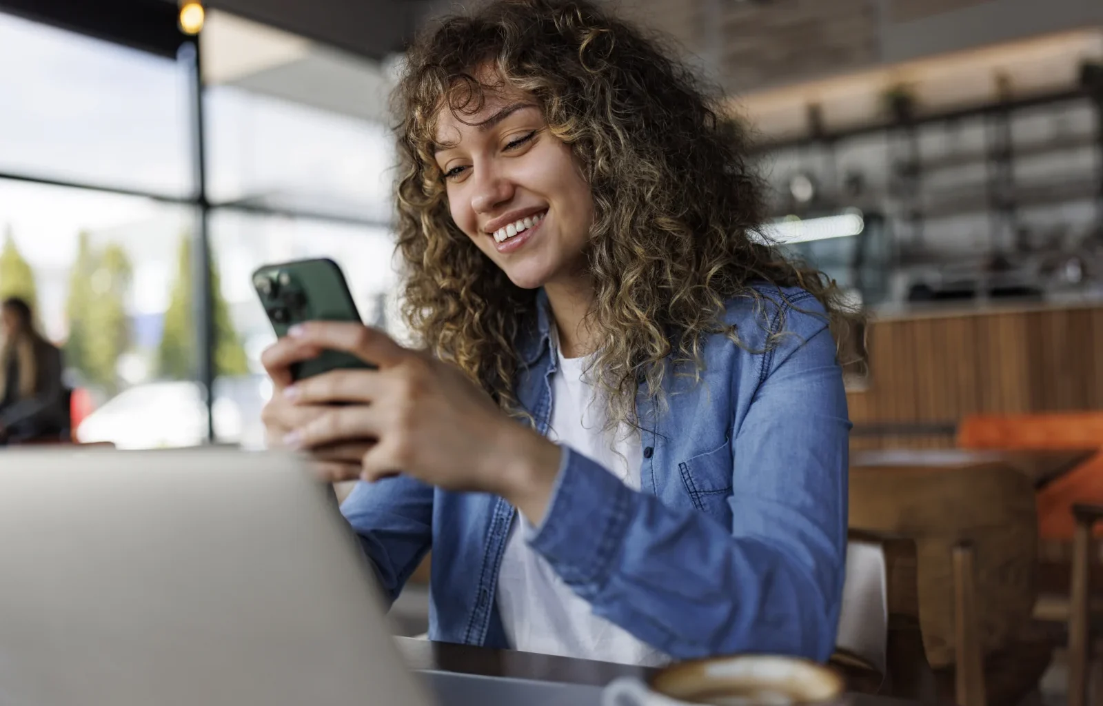 Junge lächelnde Frau mit Handy, während sie in einem Café an einem Laptop arbeitet