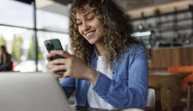 Junge lächelnde Frau mit Handy, während sie in einem Café an einem Laptop arbeitet