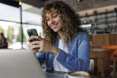 Junge lächelnde Frau mit Handy, während sie in einem Café an einem Laptop arbeitet