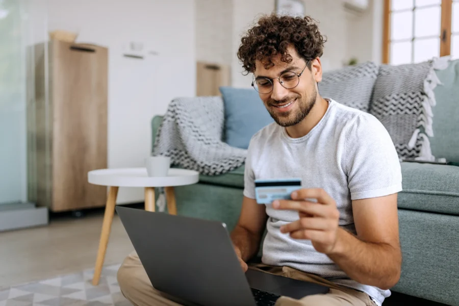 Junger lässig gekleideter Mann, der online mit Kreditkarte einkauft, während er zu Hause auf dem Boden sitzt
